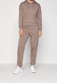 Sudadera con capucha y pantalones deportivos marrones hechos de tela suave y texturizada. Cuenta con capucha con cordón y puños. Combinados con zapatillas deportivas blancas.