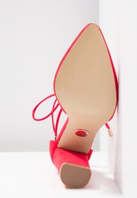 Chaussure à talons hauts en velours rouge avec un bout pointu, des brides de cheville et une semelle lisse beige avec un logo circulaire au bas.