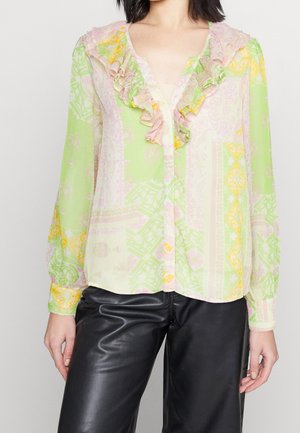 Kvinde iført gennemsigtig pastelgrøn bluse med blomsterprint og flæset V-hals samt sorte læderbukser, stående mod en ensfarvet baggrund.