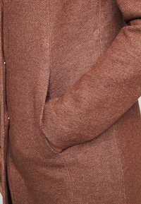 Cardigan marrone con texture, dotato di bottoni frontali e tasche laterali, che presenta un motivo in tessuto intrecciato e un aspetto morbido e caldo.