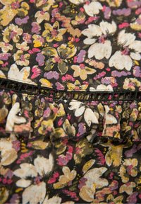 Czarny kwiatowy materiał z białymi i fioletowymi kwiatami, wyposażony w falbanę na krawędzi oraz ozdobną czarną koronkę. Całość zdobią błyszczące akcenty.