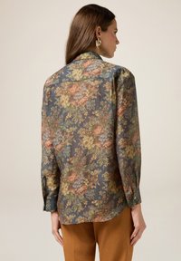 Kwiatowa bluzka z półprzezroczystego materiału, w kolorze niebiesko-szarym, z wielokolorowym nadrukiem kwiatowym, z guziczkami na mankietach i zaokrąglonym dołem.