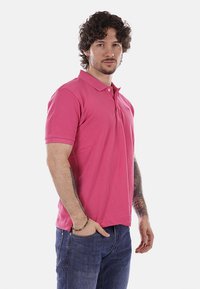 Homme aux cheveux bouclés portant un polo rose et un jean bleu, debout avec une main dans la poche et des tatouages visibles sur le bras.