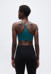 Γυναίκα με κοντά σγουρά μαλλιά, που φοράει τυρκουάζ αθλητικό τοπ με racerback και μαύρο κολάν, στέκεται με την πλάτη προς τα εμπρός σε απλό λευκό φόντο.