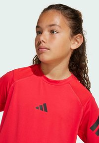 Rotes Sportshirt mit kurzen Ärmeln, versehen mit einem schwarzen Logo auf der Brust, glatter Textur und Mesh-Details an den Schultern.