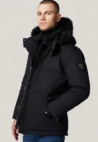 Czarna kurtka puchowa z futrzanym kapturem, zamek błyskawiczny oraz zapięcie na guziki, a także kieszeniami po bokach. Posiada naszywkę z logo na rękawie.