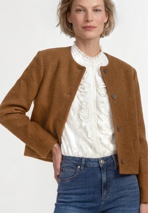 Frau trägt eine braune, geknöpfte Jacke über einer weißen, rüschigen Spitzenbluse und blauen Jeans, steht mit einer Hand in der Hüfte vor neutralem Hintergrund.