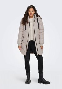 Jasnoszara pikowana kurtka z kapturem, zapinana na zamek błyskawiczny, z czarnymi akcentami, bocznymi kieszeniami i pikowanym wzorem. Noszona na kremowym swetrze i czarnych spodniach.