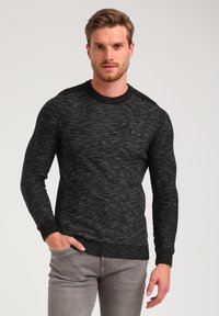 Black textured sweater with a crew neck, ribbed cuffs, and a small logo accent on the chest, paired with light gray pants.