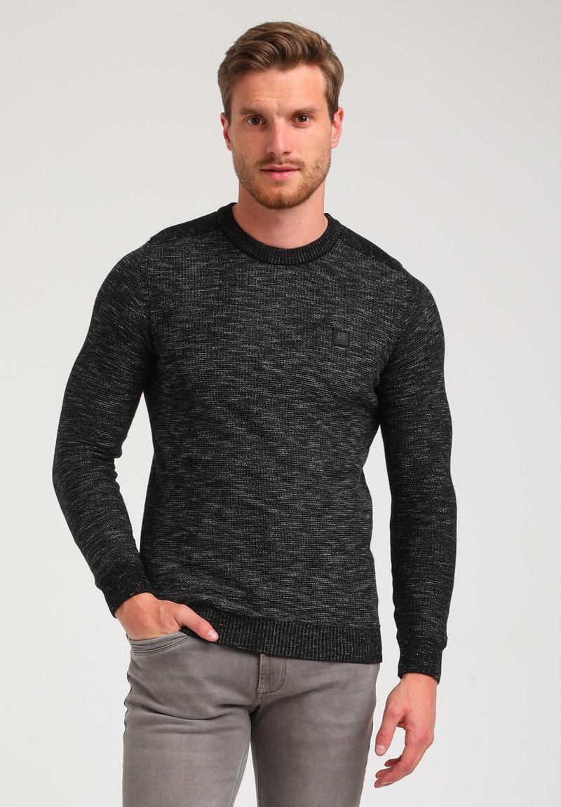Black textured sweater with a crew neck, ribbed cuffs, and a small logo accent on the chest, paired with light gray pants.