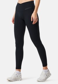 Black high-waisted leggings with a smooth texture, flat seams, and logo detailing; paired with white athletic shoes featuring a cushioned sole.
