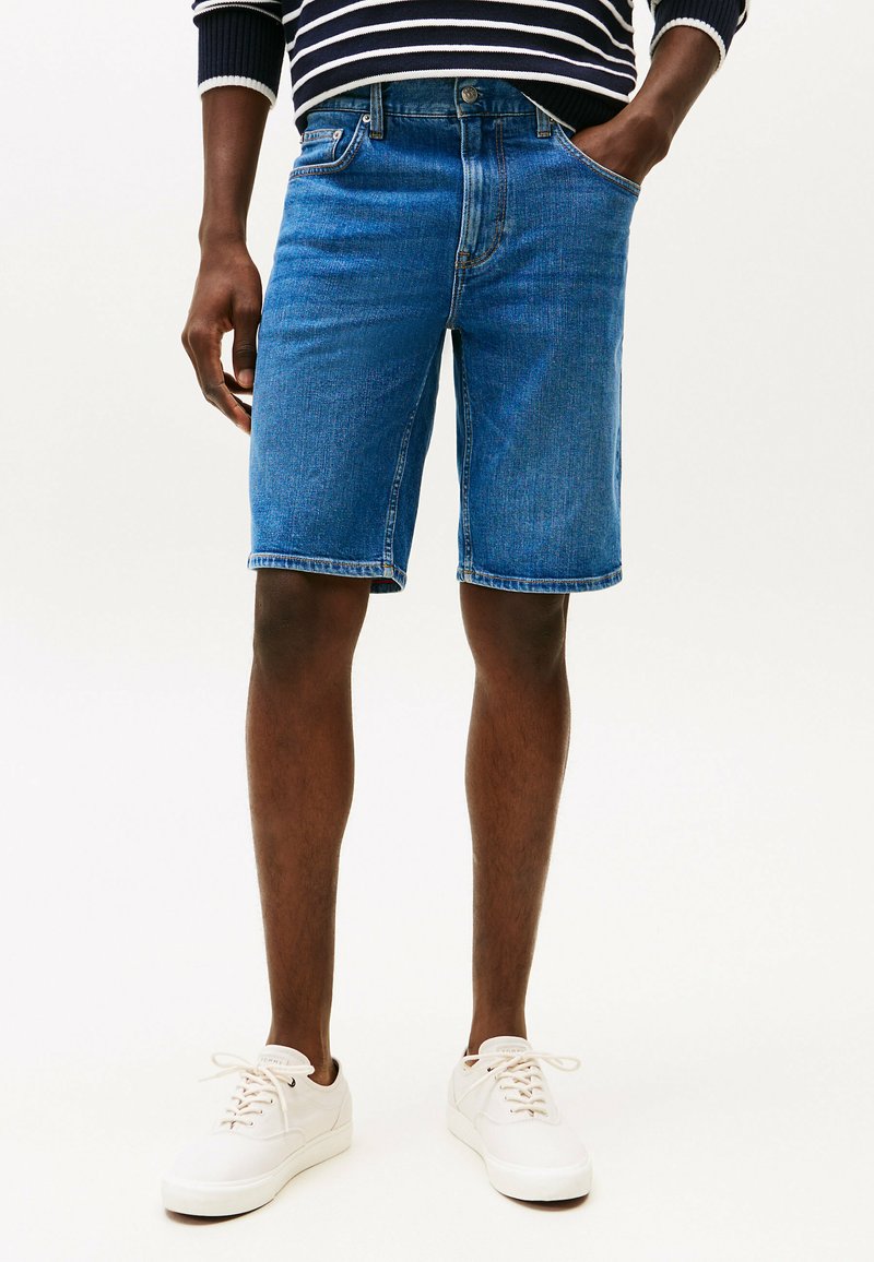 Denimshorts in medium blauw, met een klassieke vijf-pocketontwerp, een gehavende zoom en een ritssluiting met knoop. G gedragen met witte sneakers.