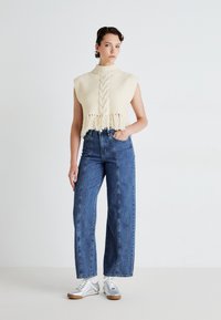 Cropped cream knit sweater with cable design and fringe detail, paired with high-waisted blue denim jeans and silver athletic shoes.
