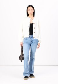 Gilet côtelé blanc avec des boutons en bois, débardeur noir, jeans évasés bleu clair, baskets noires et beiges, tenant un sac à main noir.