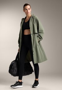 Olivgrön lätt regnjacka med huva, åtdragen midja och svarta dragkedjefickor, kombinerad med svarta leggings och sportiga skor. Svart gymväska.