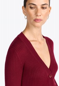 Cardigan côtelé bourgogne avec un décolleté en V prononcé et une fermeture à un bouton. Tissu texturé avec des manches longues ajustées.