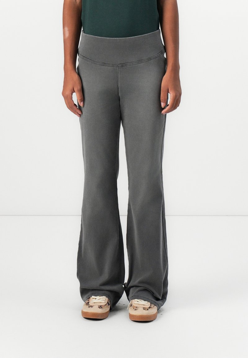 Personne portant un pantalon gris évasé et des baskets beiges, debout devant un fond blanc uni, mains le long du corps, seule la partie inférieure du corps est visible.