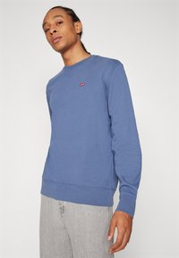 Blauwe crewneck sweatshirt van zachte stof. Heeft een klein rood logo op de borst en ribgebreide manchetten. Casual, relaxte pasvorm.