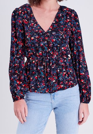 Femme portant un chemisier à manches longues à fleurs rouges, bleues et blanches et un jean bleu clair sur un fond uni.