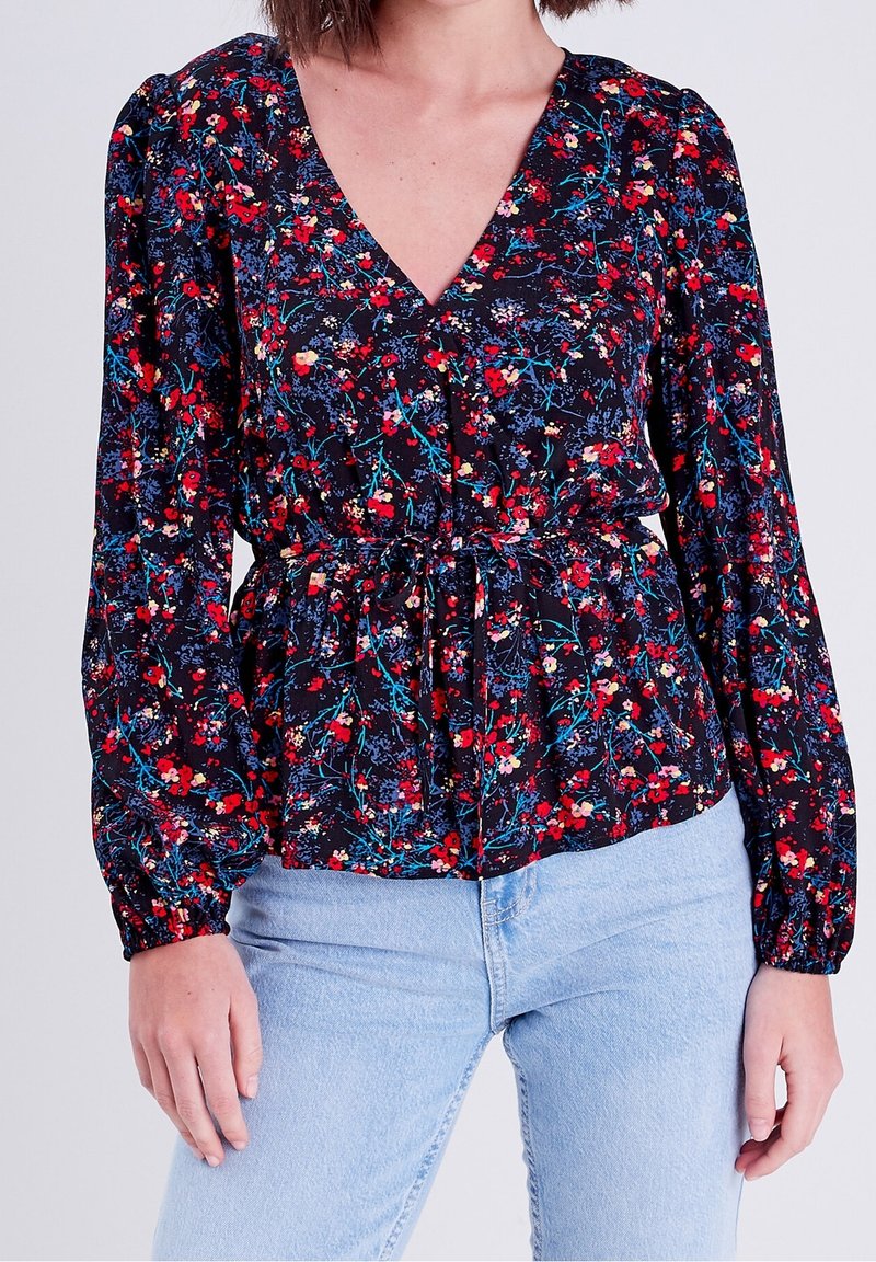 Femme portant un chemisier à manches longues à fleurs rouges, bleues et blanches et un jean bleu clair sur un fond uni.