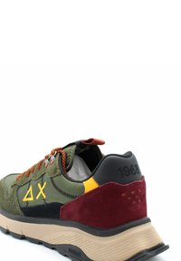 Sneaker verdi e neri con accenti bordeaux e gialli, dotati di una parte superiore in tessuto testurizzato, suola in gomma e lacci arancioni. Etichette e loghi visibili.