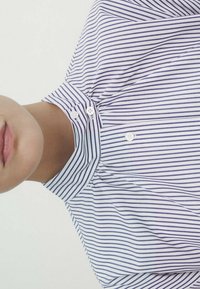 Chemise à boutons avec des rayures violettes et blanches, en tissu de coton doux, col haut et deux boutons à l'avant. Coupe ajustée avec un léger détail froncé.