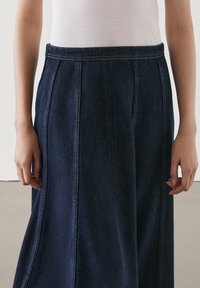 Mörk denim långkjol med hög midja, sidofållar och kontrasterande sömmar. Tyget har en slät yta och en strukturerad form.