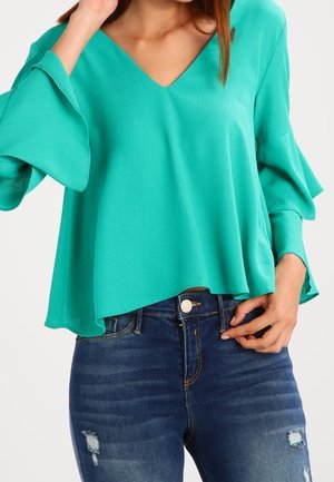 Nainen, joka käyttää löysää sinivihreää pitkähihnaista puseroa, jossa on levenevät kalvosimet, ja tummansiniset kulutetut farkut.