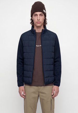 Jeune homme portant un bonnet marron, une veste matelassée bleu marine, une chemise marron et un pantalon cargo beige, debout devant un fond blanc uni.