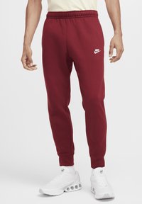 Red sweatpants with an elastic waistband, cuffed ankles, and a small white Nike logo. Paired with white shoes featuring a textured pattern.