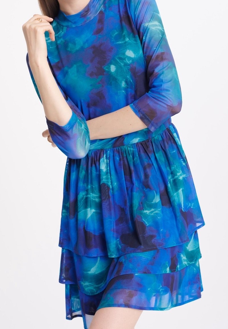 Robe aux couches bleu et sarcelle avec un col montant, manches longues et une silhouette fluide, présentant un motif semblable à une aquarelle et un tissu transparent.