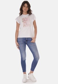 T-shirt bianco a maniche corte con stampa floreale e dettagli rosa, abbinato a jeans skinny blu e sneaker bianche.