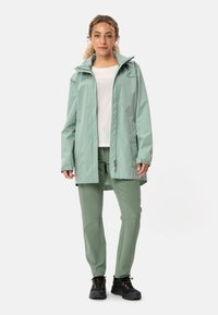 Giacca impermeabile verde chiaro con cappuccio, chiusura con zip e tasche, abbinata a pantaloni verde chiaro e scarponi da trekking neri.