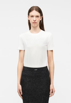 Ženska s ravnimi rjavimi lasmi nosi bel kratkoročni top in črno teksturirano krilo, stoječo pred enostavnim svetlim ozadjem.