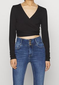 Svart långärmad omslagstopp kombinerad med högmidjade blå denimjeans som har tre guldknappar vid midjan och sidofickor.