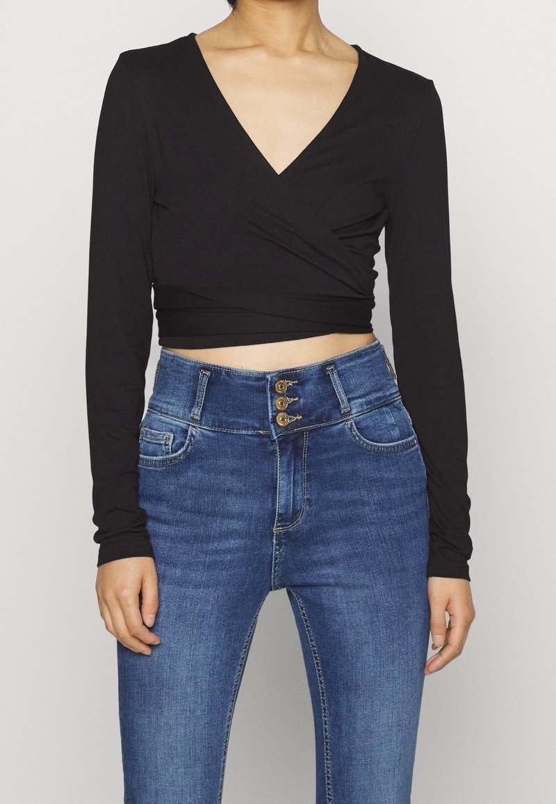 Svart långärmad omslagstopp kombinerad med högmidjade blå denimjeans som har tre guldknappar vid midjan och sidofickor.