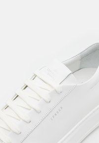 Witte leren sneaker met platte witte veters, een gladde textuur en een reliëflogo op de tong en zijkant. Minimalistisch ontwerp en een laag profiel.
