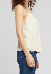 Cami top in light beige satin fabric, featuring thin straps, a relaxed fit, and a smooth, glossy texture. Styled with blue jeans.