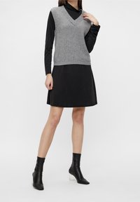 Frau in einem grauen ärmellosen Strickwesten über einem schwarzen Langarmkleid und schwarzen Stiefeletten mit transparenten Absätzen vor einem weißen Hintergrund.