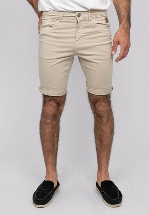 Homme portant un short beige retroussé, associé à un haut blanc et des chaussures noires sans lacets, debout devant un fond uni.