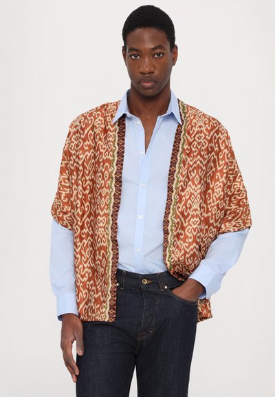 Kimono estampado en tonos cálidos de naranja y crema, con diseños intrincados y un ribete en contraste. Lleva puesto encima de una camisa azul clara y jeans oscuros.