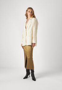 Blazer de textura crema con bordes deshilachados, combinado con una falda dorada de seda con una abertura lateral y botas negras de punta afilada hasta la rodilla.
