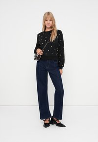 Sweatshirt noir avec des petits motifs en cœur doré, associé à un jean bleu foncé à jambes larges. Les accessoires incluent un petit sac à bandoulière noir et des chaussures.