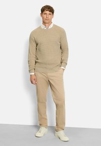 Beige gestrickter Pullover über einem hellblauen Hemd mit Kragen, kombiniert mit beigen Chinos und weißen Sneakers, vor einem einfarbigen Hintergrund stehend.