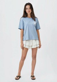 Svetle modré tričko s krátkým rukávem a uvolněným střihem, které je vrstveno přes bílé šortky s volánky, doplněné černými sandály.