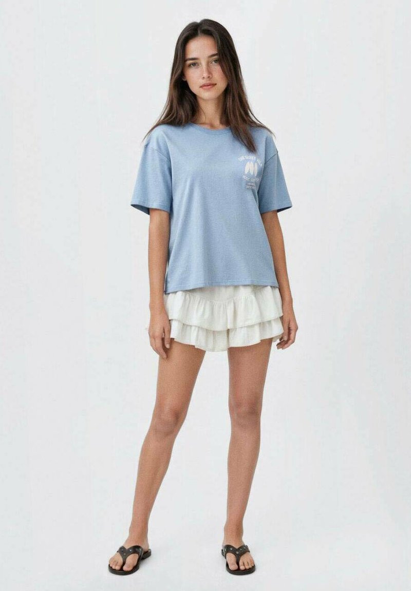 Svetle modré tričko s krátkým rukávem a uvolněným střihem, které je vrstveno přes bílé šortky s volánky, doplněné černými sandály.