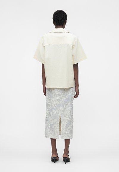 Άτομο που στέκεται με την πλάτη, φοράει μια εκρού κοντομάνικη μπλούζα, υφασμάτινη φούστα μέχρι το γόνατο με σκίσιμο στο πίσω μέρος και μαύρα σανδάλια με τακούνι.