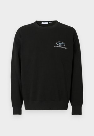 Melnā kokvilnas džemperis ar apaļu kaklu, garām piedurknēm un ribinātām rokdarbēm. Uz krūšu daļas ir balts izšūts logo.