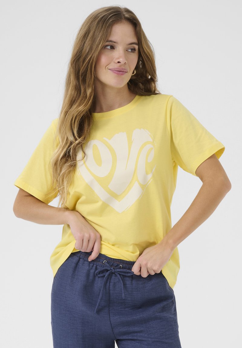 Kollane puuvillane T-särk lühikeste varrukatega, millel on valge südamekujuline "love" graafika. Kanda siniste nöörpükste kõrval.