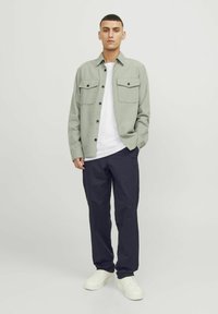Lys grøn skjorte med knapper og to brystlommer, hvid t-shirt under, navy bukser og hvide sneakers; minimalistisk design, afslappet stil.